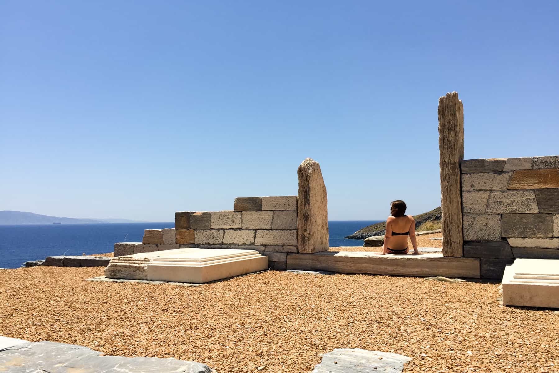 A woman sits in the doorway of the ruins of an ancient temple monument in Kea island, looking out to the dark blue sea