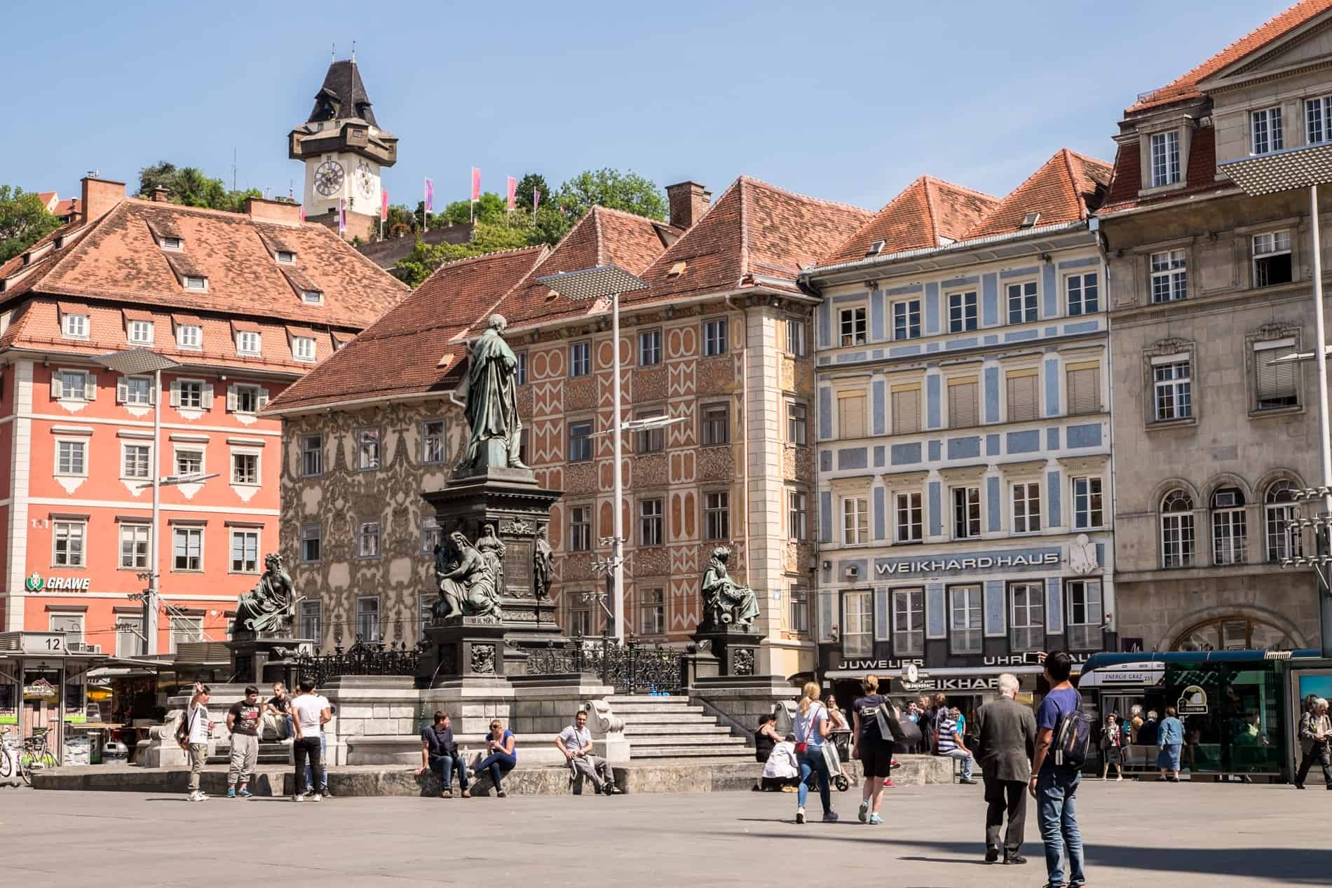 The old town square of Graz Hauptplatz with statues, coloured buildings and a view to the clock tower on the hill. 