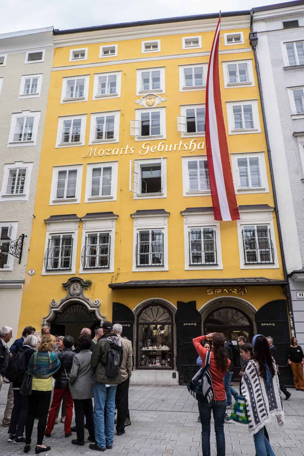 People stand outside a tall, rectangular yellow house with a red and white stripped Austrian flag. The words on the building read: "Mozart's Geburtshaus" (Mozart's Birth House). 
