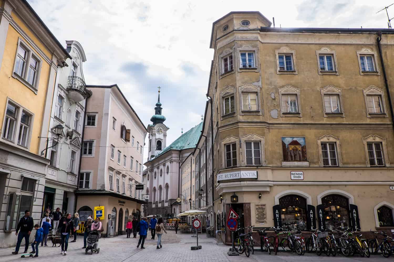 View of the Andräviertel Neighbourhood in Salzburg comprised on yellow and pink pastel coloured buildings, mint green rooftops and spires and a worn, golden brown four story building with the sign, "StRupert Antik". 