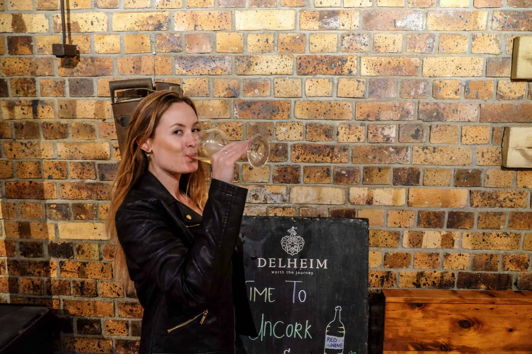 A woman in a black jacket drinks a glass of wine in front of an exposed brick wall. The blackboard sign says 'Time to Uncork' with the logo of Delheim Wine Estate in Stellenbosch