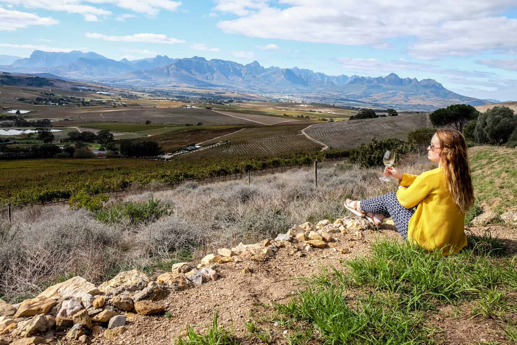 A woman wearing a yellow cardigan sits on a rocky hillside, holding a glass of wine. She looks out over the vast patchwork green and brown landscape of the Jordan Wine Estate in Stellenbosch, South Africa 