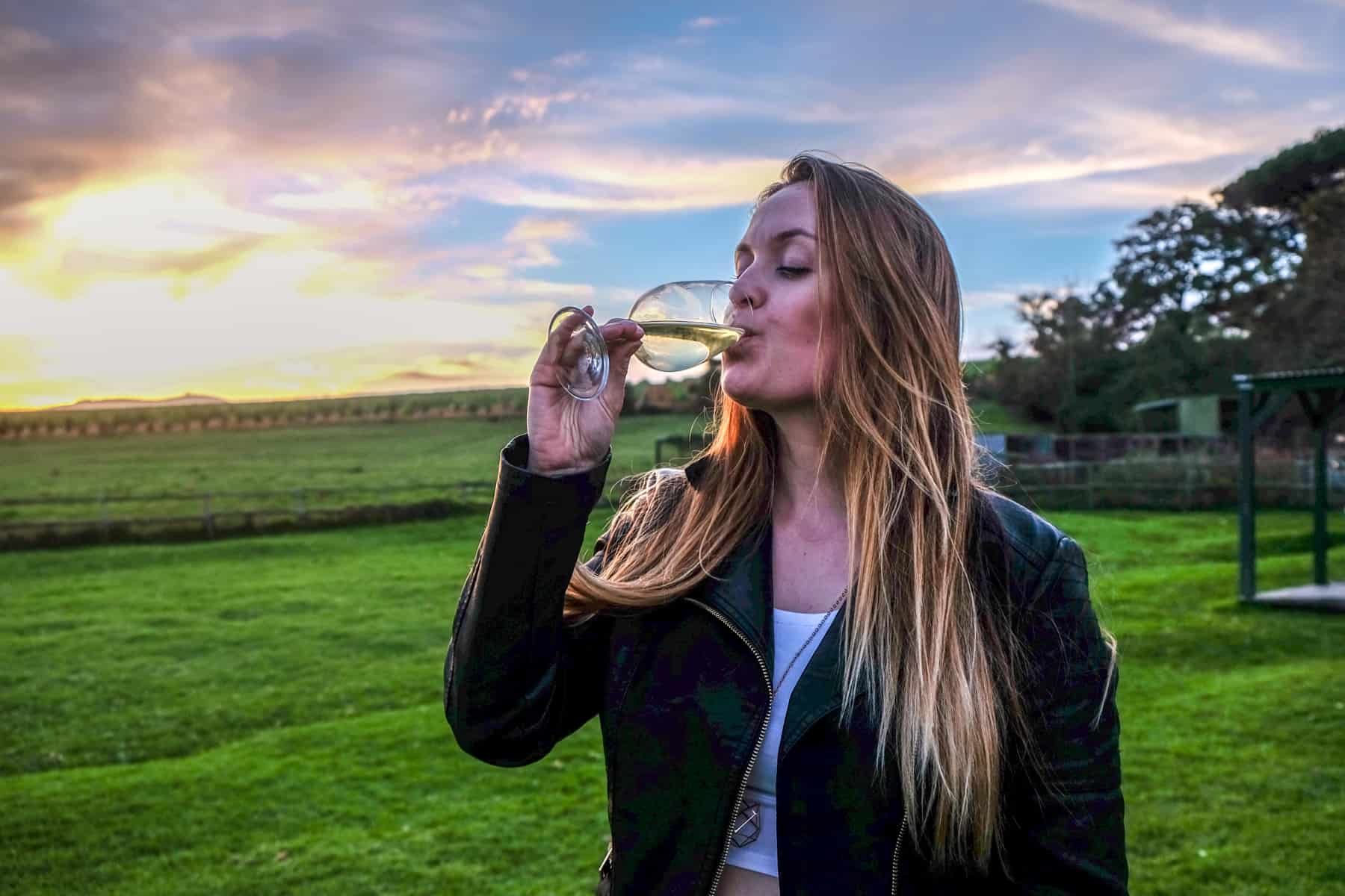 A woman with long hair and wearing a black jacket drinks wine in the green field of a wine state in Stellenbosch, under the pinks, blue and orange sunset sky.