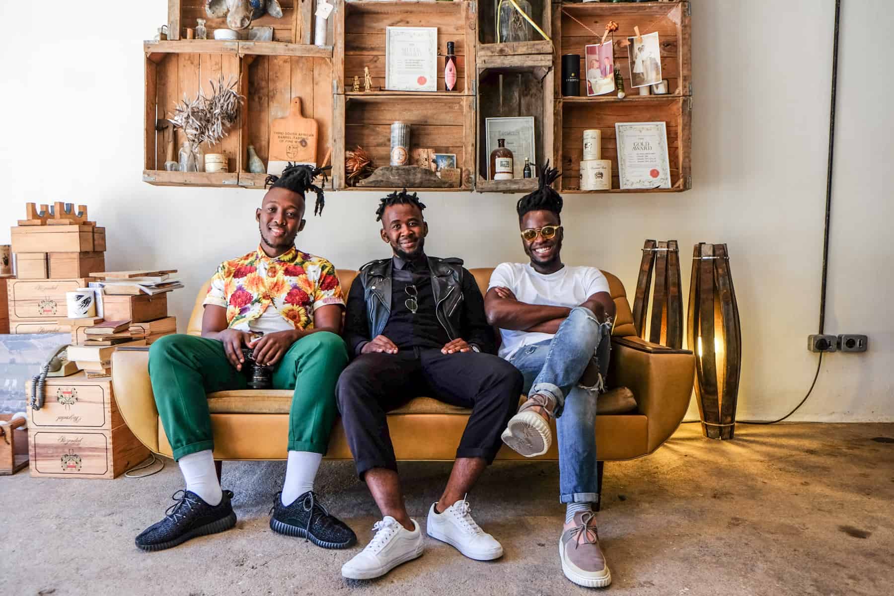 Three men sit on a yellow sofa, backed by a wall mounted wooden shelving frame containing certificates and bottles of vinegar on display