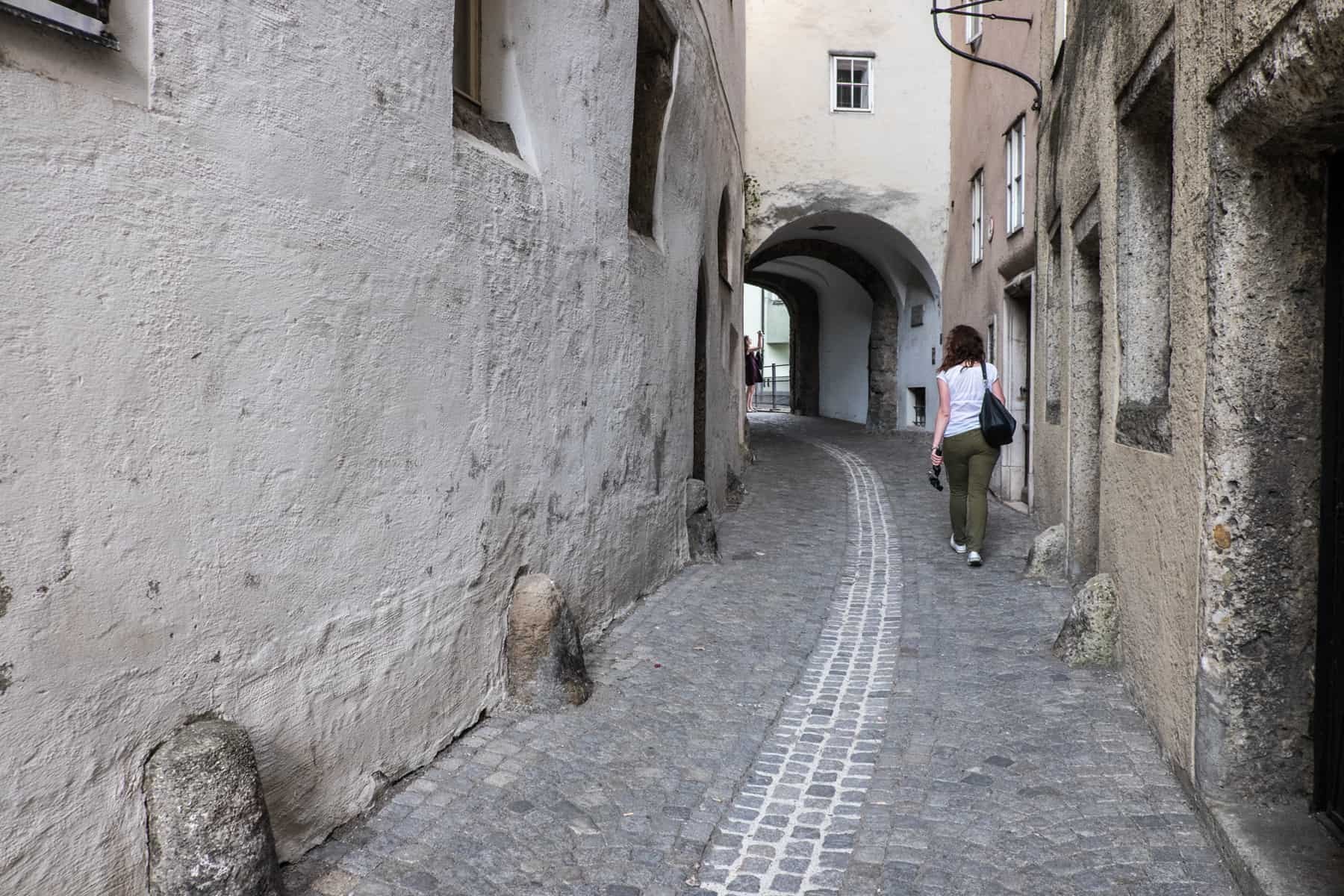 A woman in a white t-shirt and green trousers walking in a narrow cobblestoned alleyway towards a white wall arch in old Salzburg, Austria. 