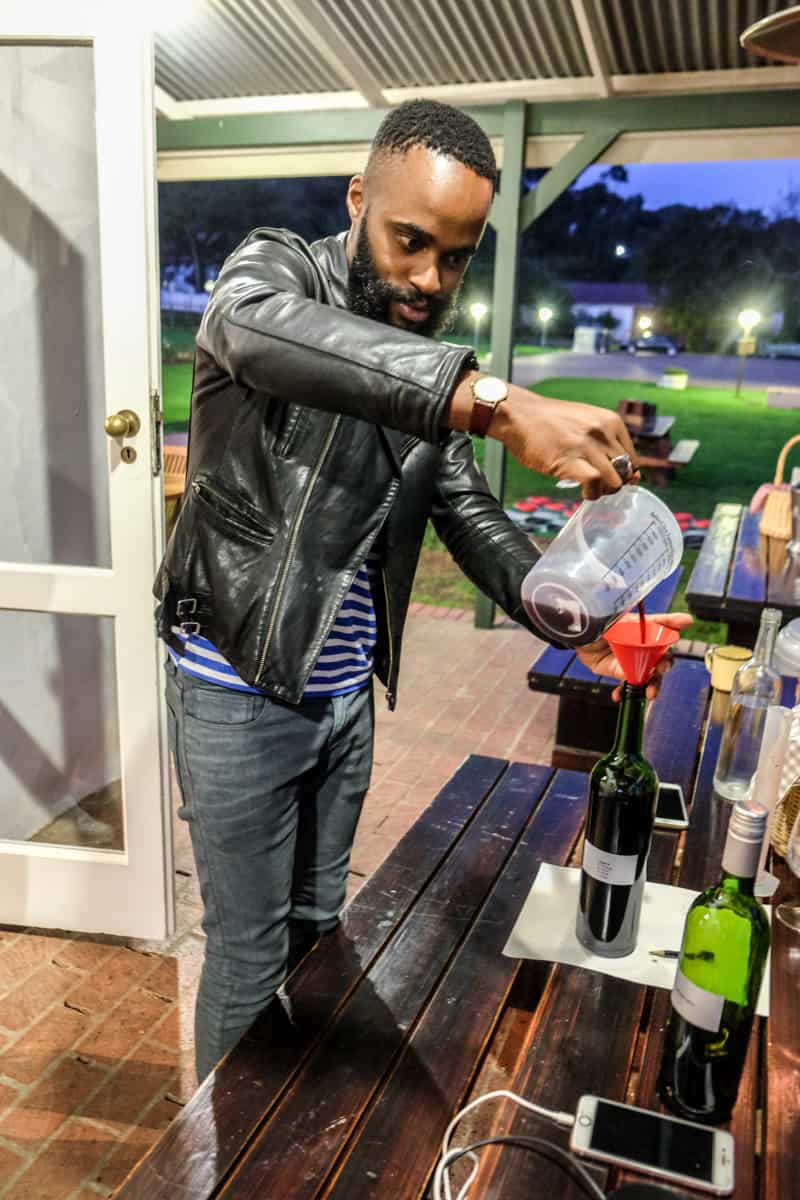 A man standing at a table pours a wine mixture into a wine bottle using a plastic measuring jug, as part of a wine making class in Stellenbosch