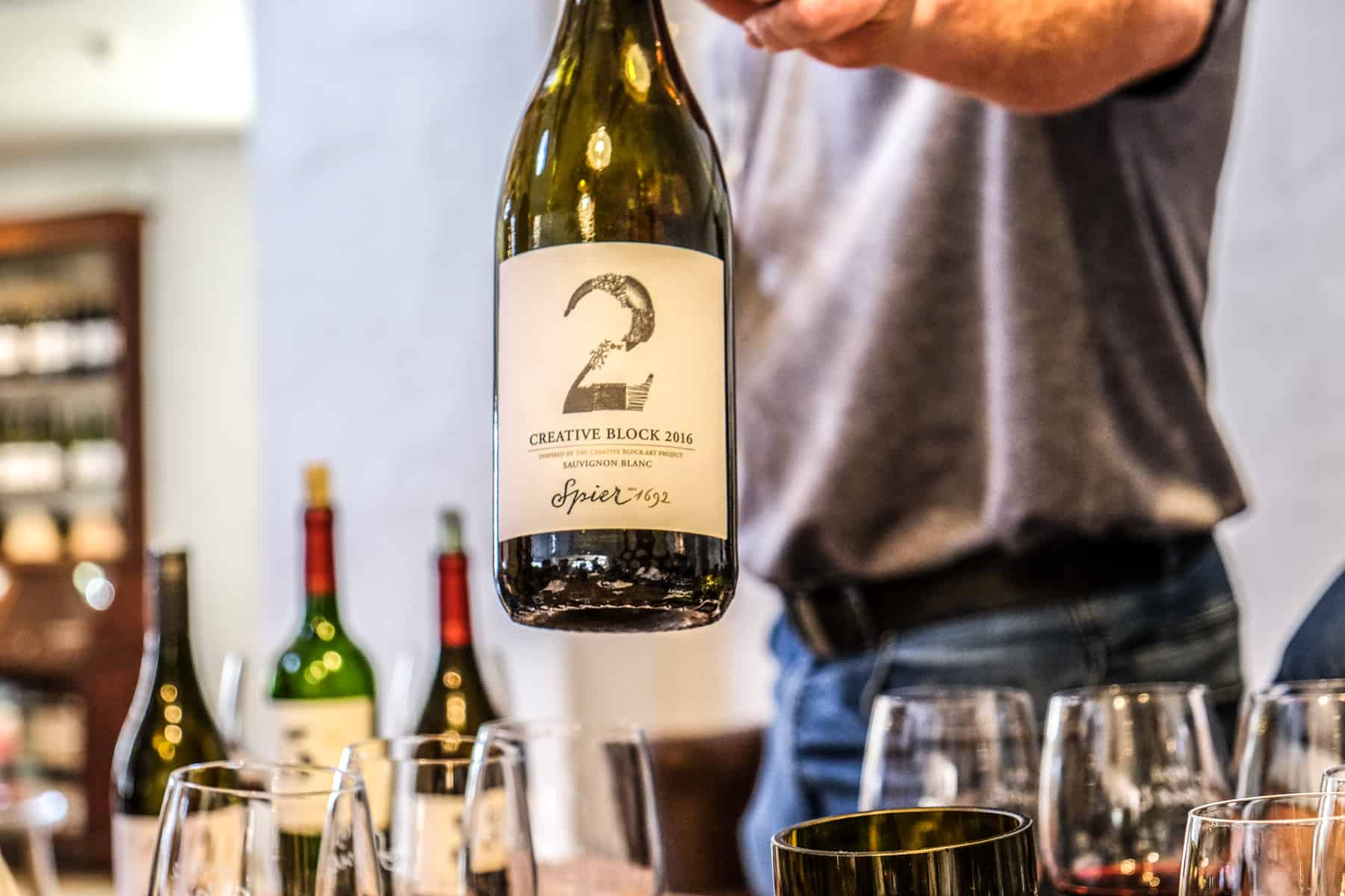 A man in grey t-shirt holds out a bottle of wine for review. The beige label features an illustrated number 2 and the words 'Creative Block 2016. Sauvignon Blanc. Spier 1962'. 