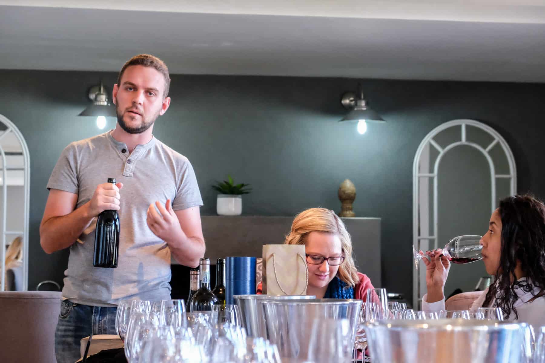 A man in a grey shirt holds a bottle of wine while explaining during a tasting. A woman next to him looks down, while another tries a red wine. The table in front is full of empty wine glasses