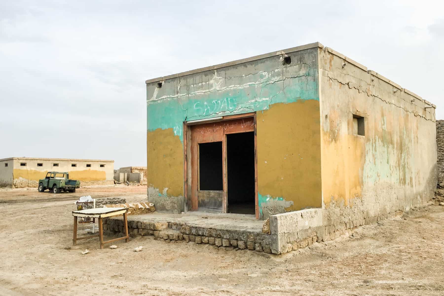 A low, square building painted in mint green and yellow is left abandoned and fading. A wooden table has been left outside the building.