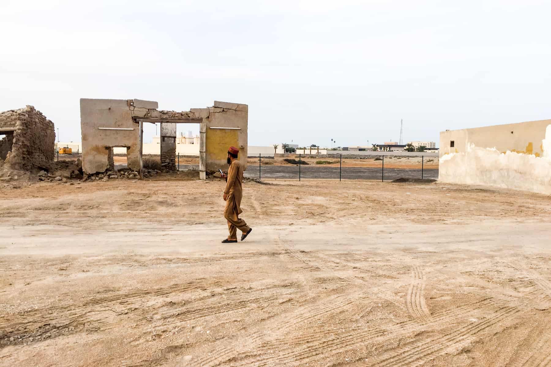 A man in dark beige overalls and a red hate walks through a sandy area in front of crumbling buildings that make up a ghost town in Ras Al Khaimah, UAE
