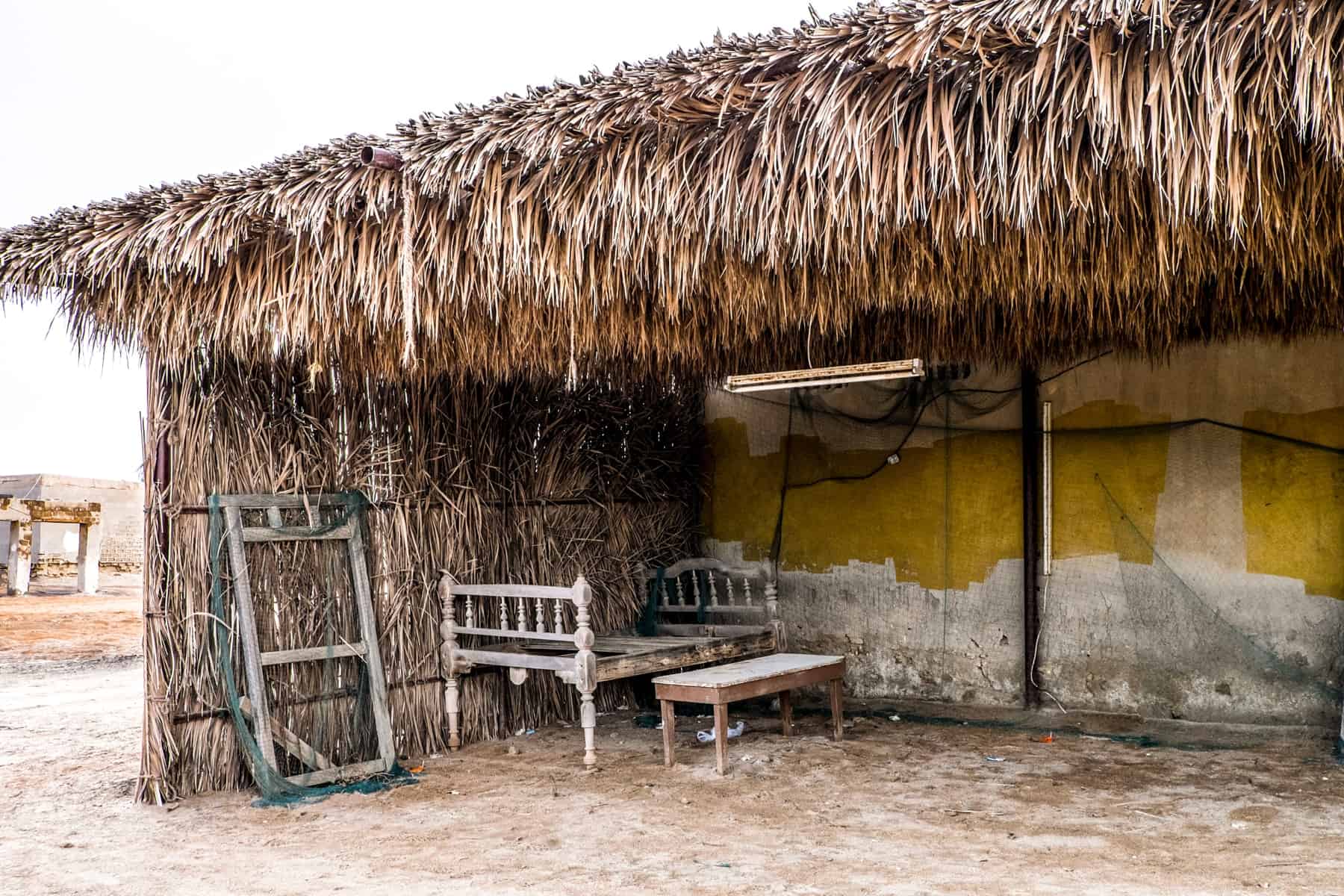 An abandoned house whose roof and one wall is made of dried reeds, contains an abandoned bed frame and table - remnants of the ghost town in Ras Al Khaimah 