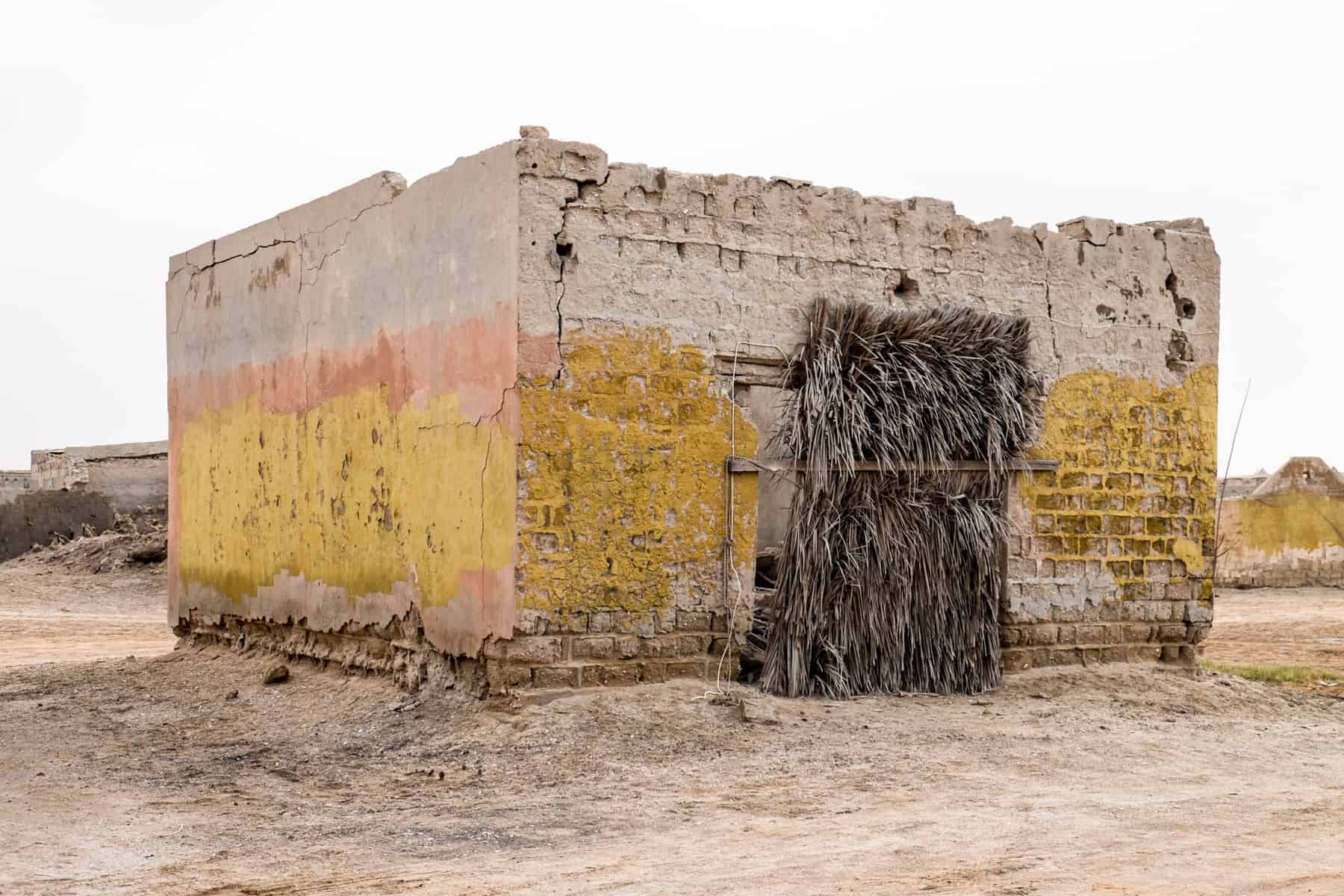 A low rectangular building with no roof, whose bottom half is painted in yellow and with a reed filled doorway, stands solitary and abandoned in Ras Al Khaimah's ghost town