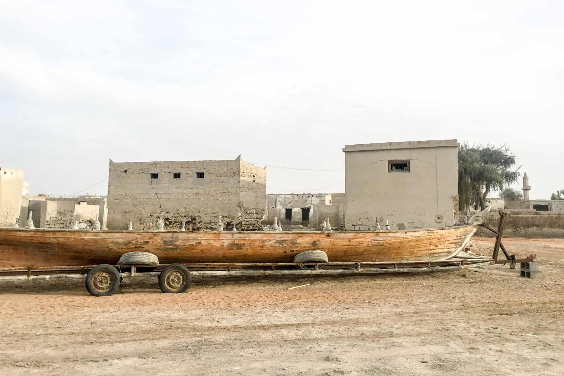 A long, light wooden boat, left abandoned in the orange coated ghost town of Al Jazirat Al Hamra in Ras Al Khaimah. Two deserted block buildings can be seen in the distance. 
