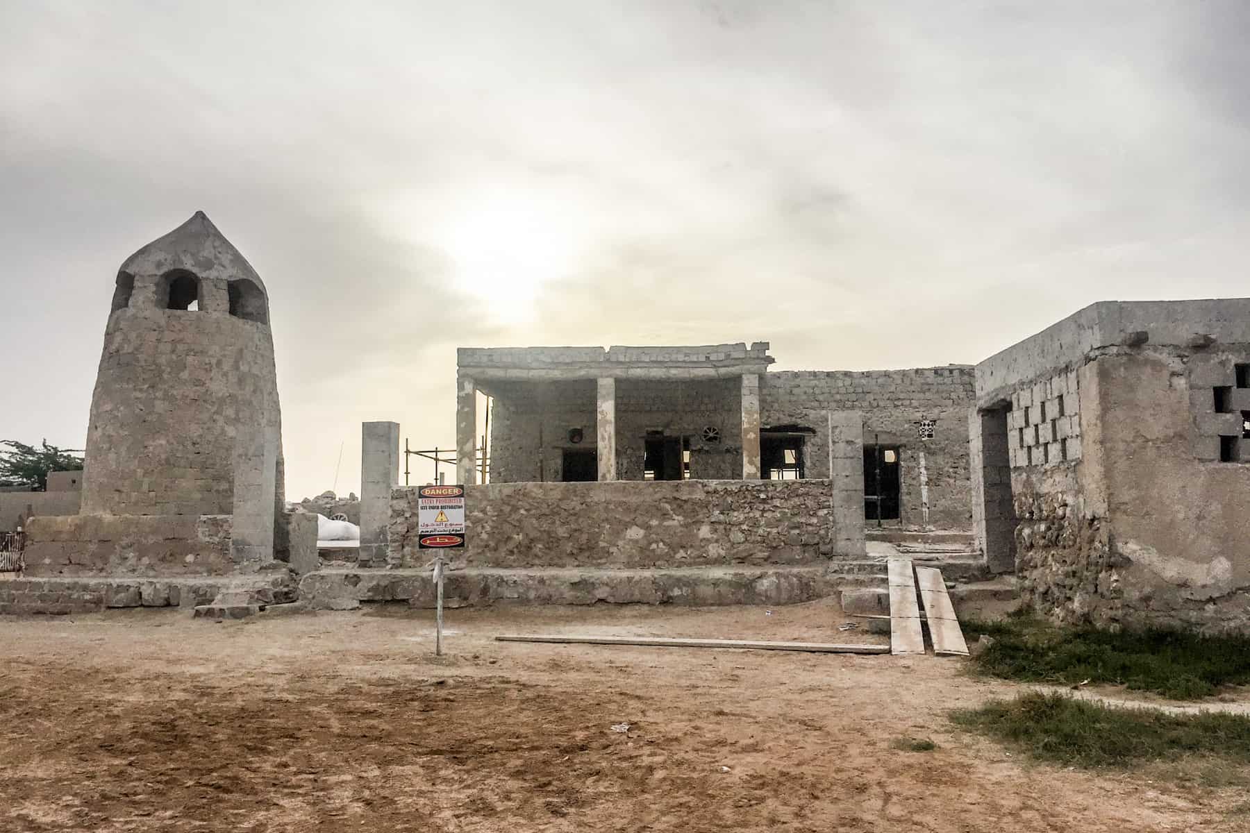 A grey tower and low building structures, abandoned and unfinished in an orange sandy area 