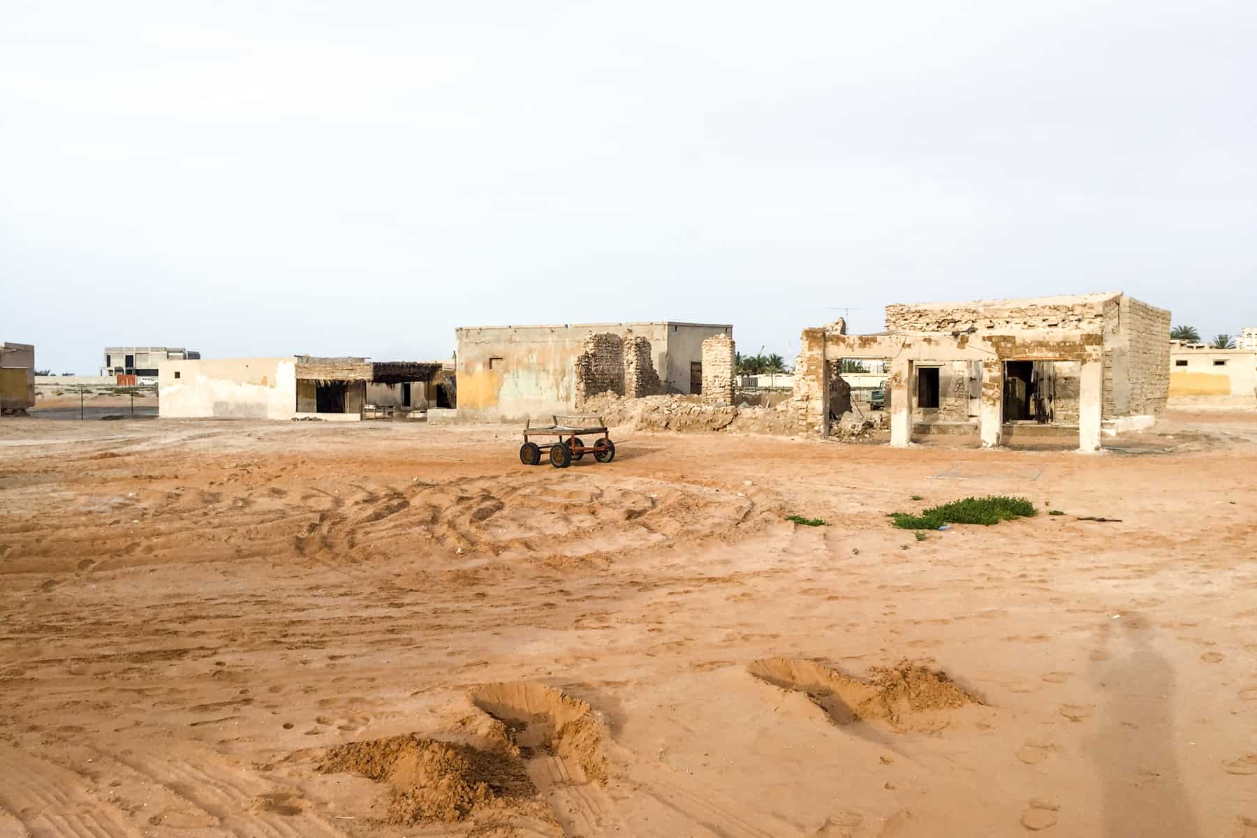 A flat buggy car stands alone in the orange sand, in front of crumbling rectangular buildings in Al Jazirat Al Hamra ghost town in Ras Al Khaimah 