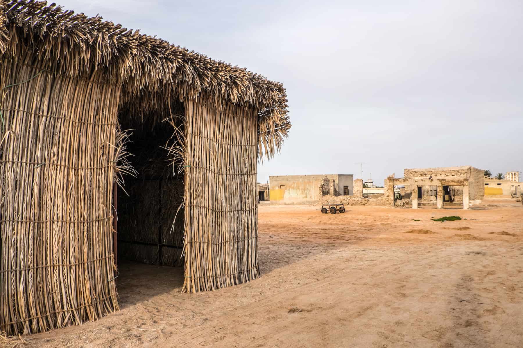 A box shaped building made of reeds and with no door, stands abandoned on orange sand. Other abandoned stone structures can be seen in the background. 