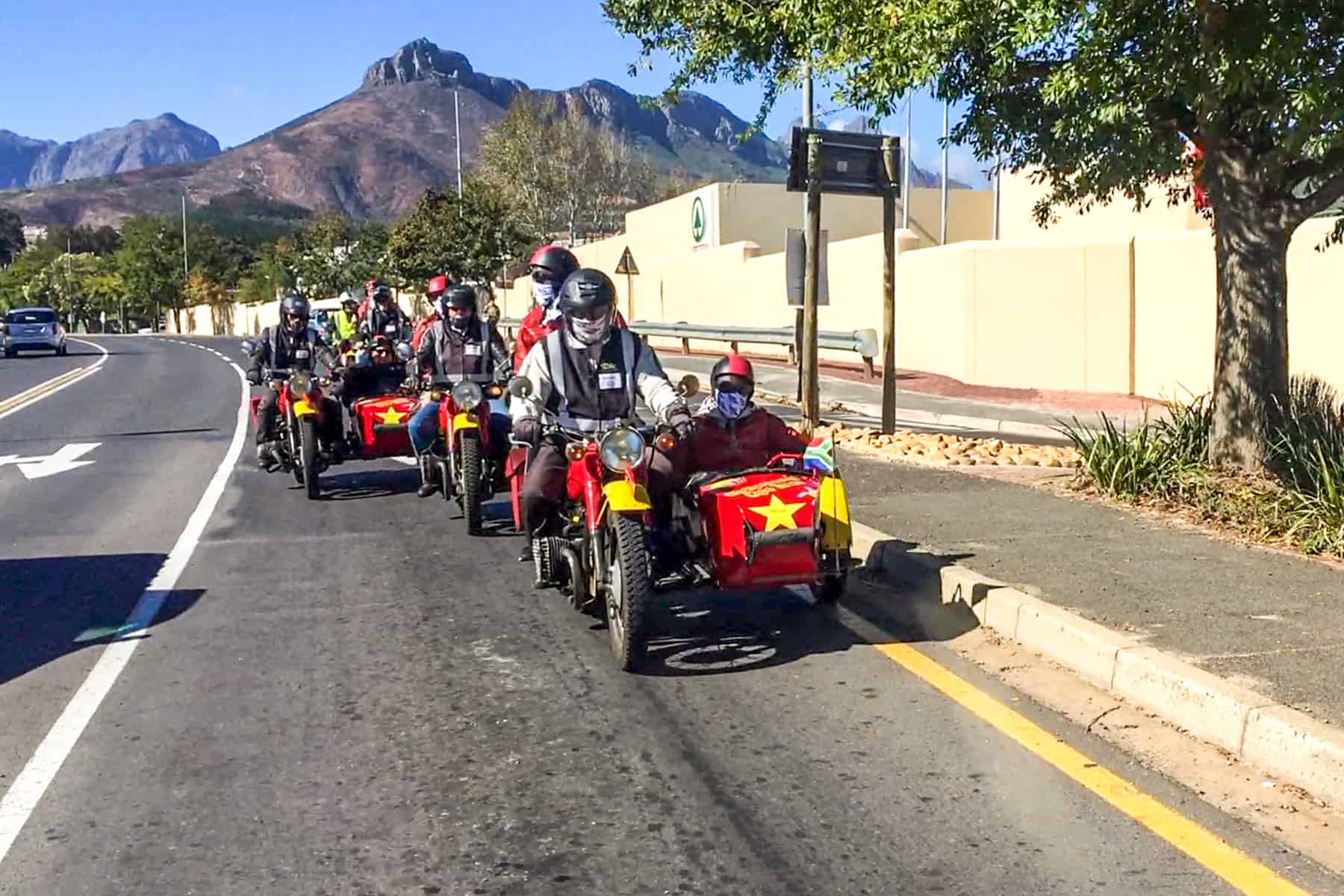 A group of people riding in red sidecars, attached to red motorbikes on a road through Stellenbosch, South Africa on a Cape Town day trip.