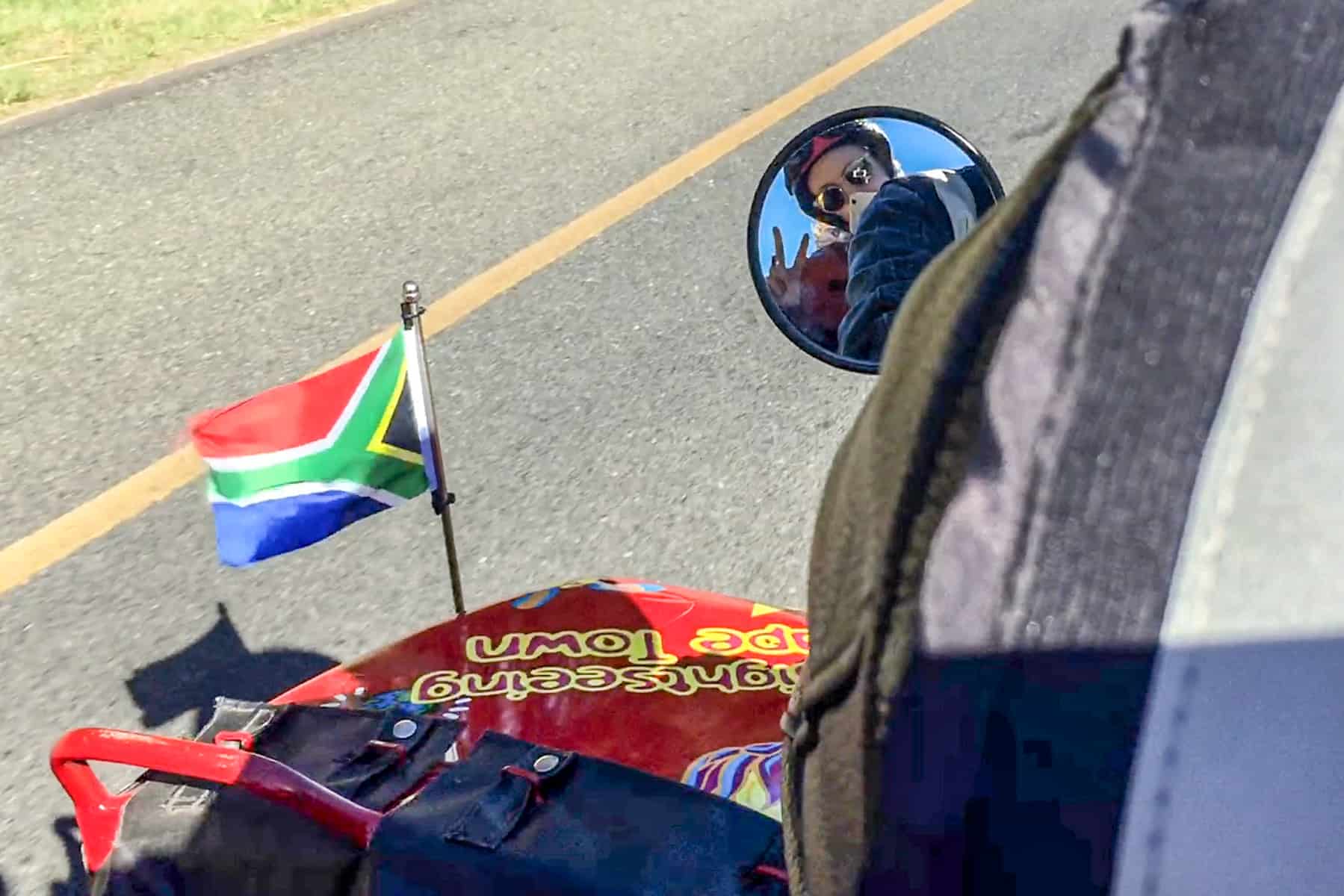 A woman looks into the mirror of a motorbike. Next to her is the red detail of the sidecar reading "Cape Town Sidecars" and a small South African flag waving in the wind.