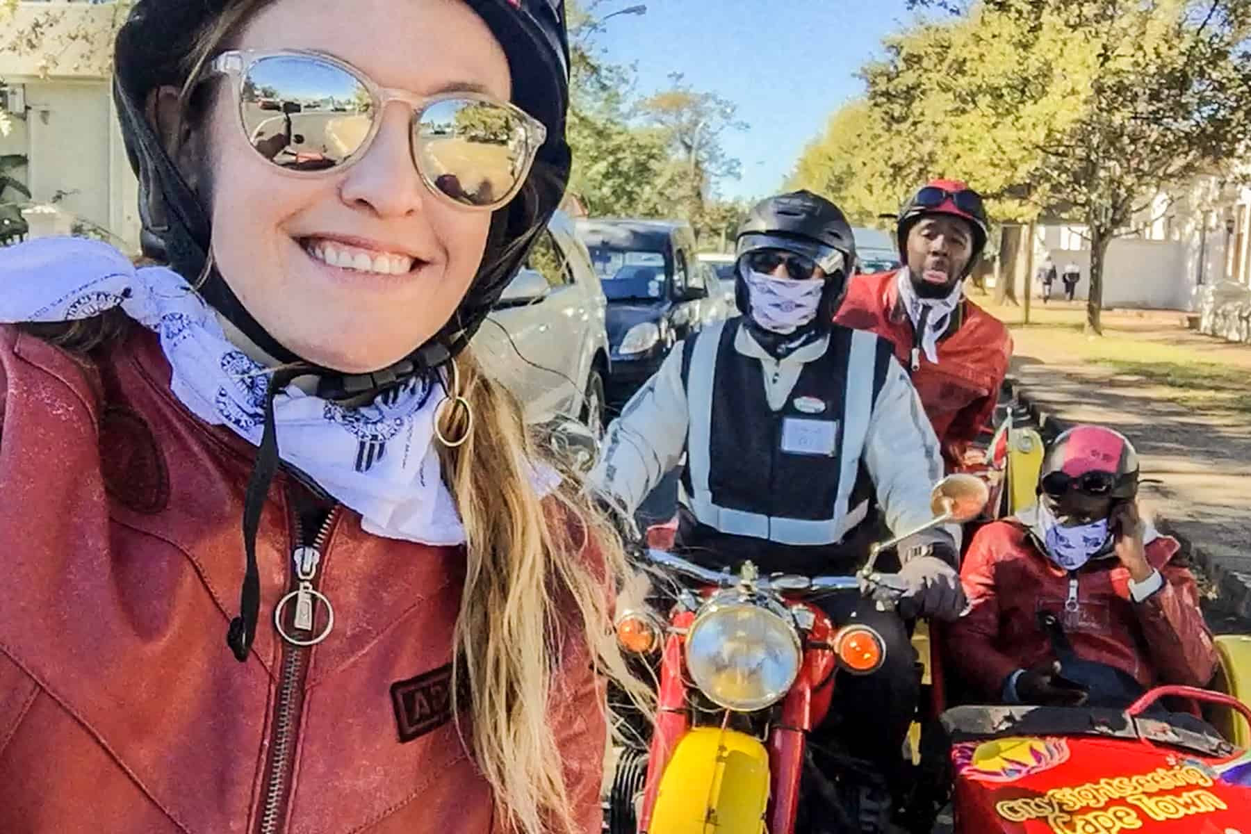 A woman wearing a red leather jacket takes a selfie. Behind her a group of other passengers, and a driver wearing white, look towards the camera from their red sidecars.