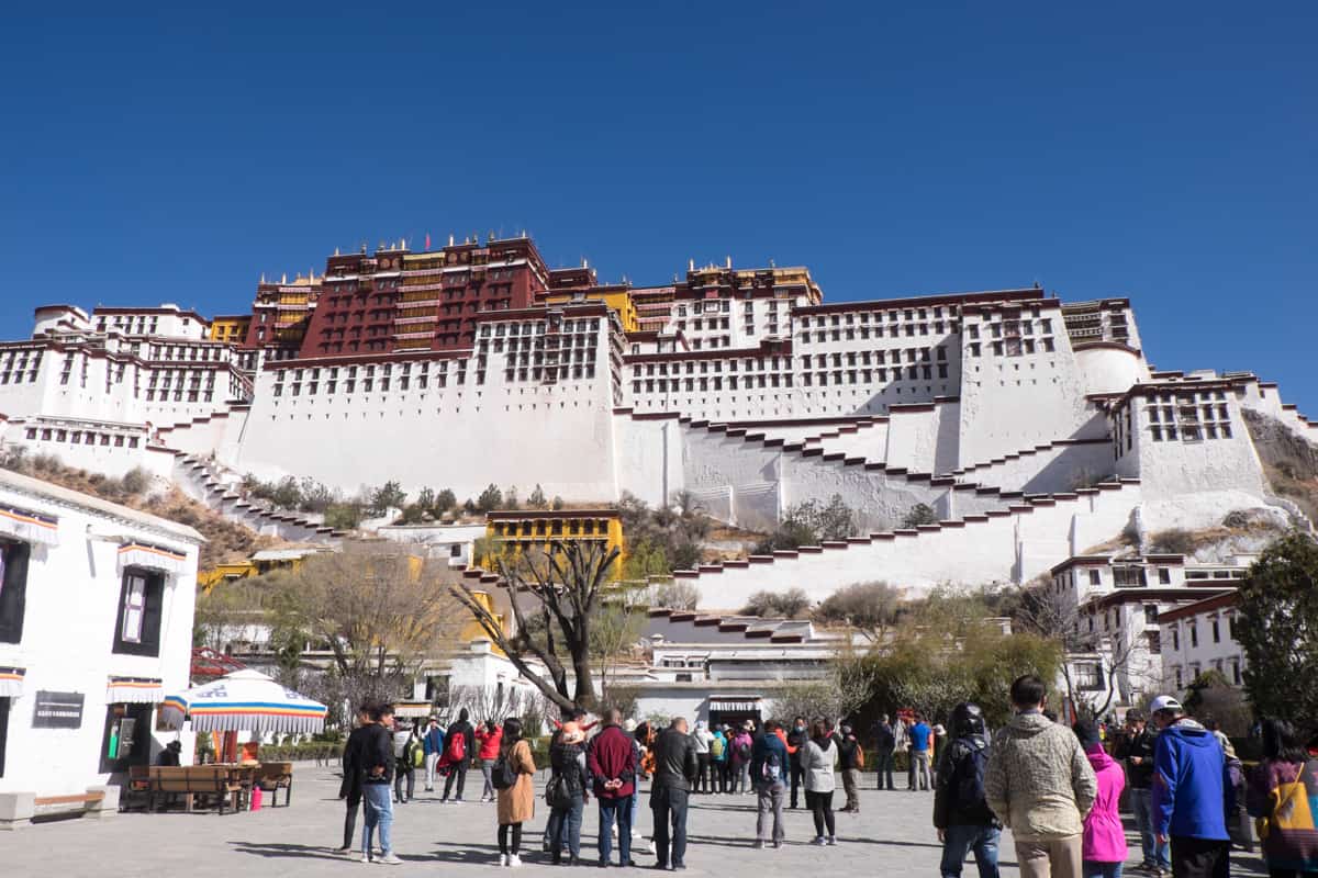 People approaching the white exterior of the hilltop Potala Palace in Lhasa, Tibet, with ochre red coloured upper buildings and yellow lower level buildings