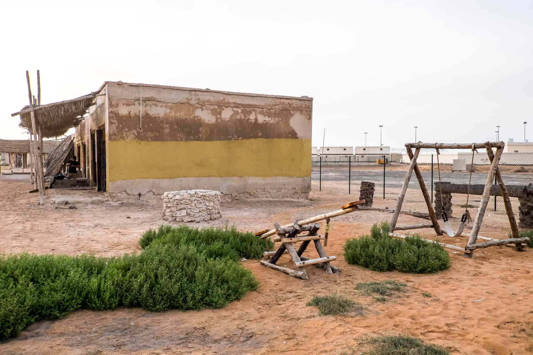 A abandoned playground, with a wooden swing and sea-saw, remains on the orange sand next to a yellow painted abandoned building in Ras Al Khaimah. 
