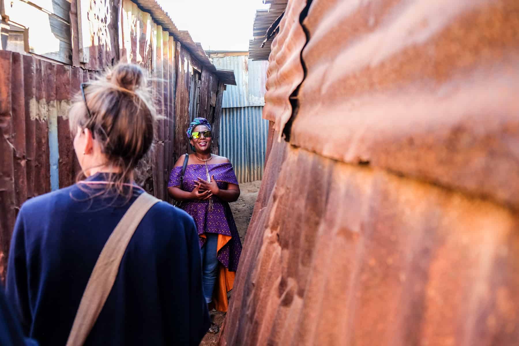 A woman in a purple dress and blue head covering looks towards the camera while standing in a narrow gap between rusting iron house walls in a township in South Africa. A woman in a blue cardigan walks towards her while on a tour.