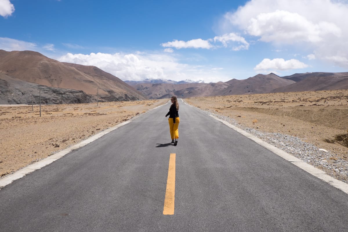 Woman in yellow pants walking on a wide and long road in Tibet, with dry land on either side and mountains in the background