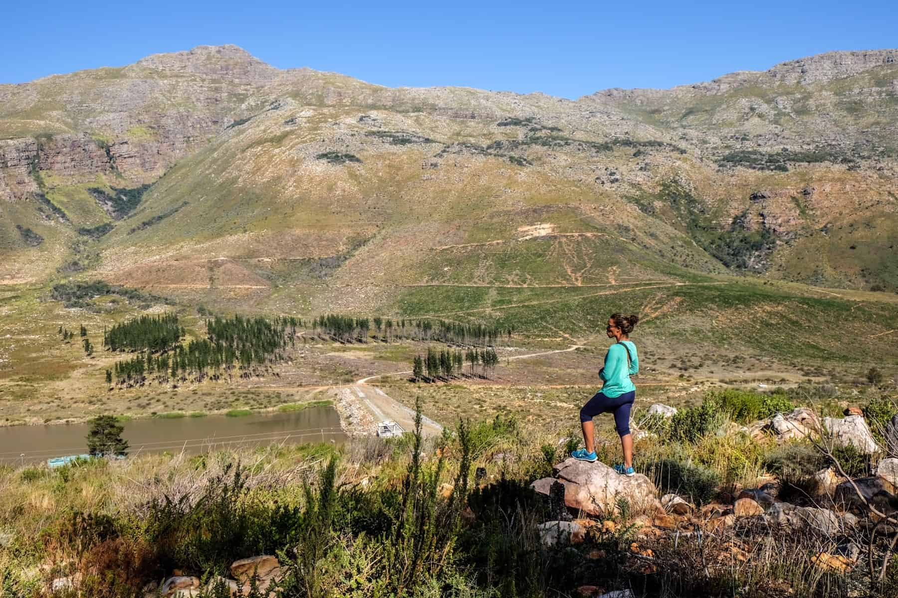 A woman wearing green stands on a rock in the middle of a vast and open green area backed by silvery, rocky hills. A small section of Jonkershoek Nature Reserve in the Western Cape.