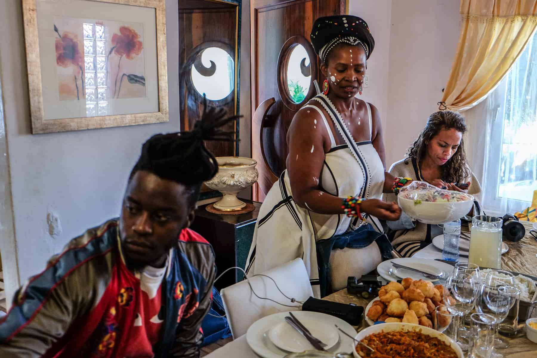 A woman wearing a white dress serves a bowl of food at a dibber spread in her home in Kayamandi township in South Africa. A man sits on the left and a woman to the right, with more bowls of food in front of them 
