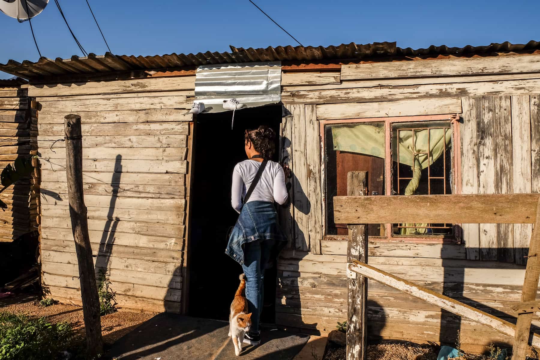 A woman in grey top and blue jeans walks into a wooden hut - a typical house in a township in South Africa. A white and ginger cat passes by her feet. 