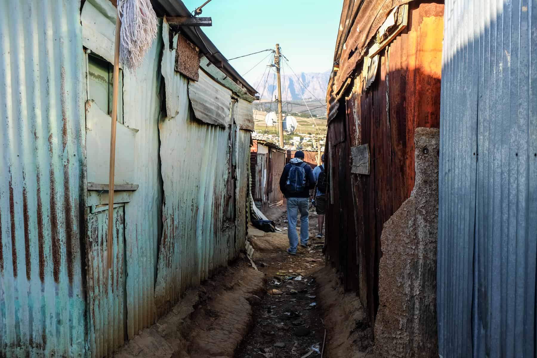 A man with a backpack walks down a narrow, dirt track between rows of light green and blue painted metal houses in the Kayamandi township in South Africa