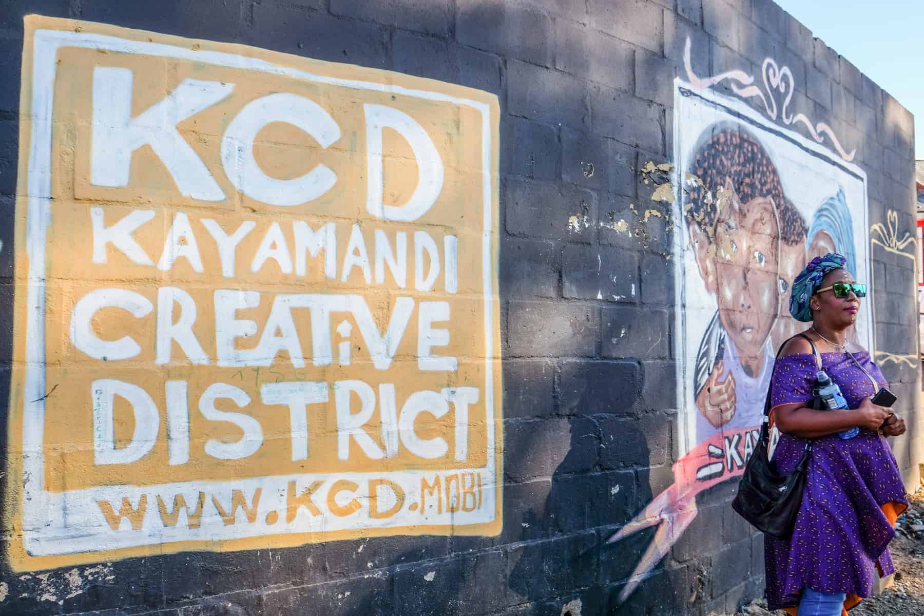 A woman in purple dress, a local guide at the Kayamandi Township in South Africa standing in front of street art as a sign for the KCD Kayamandi Creative District