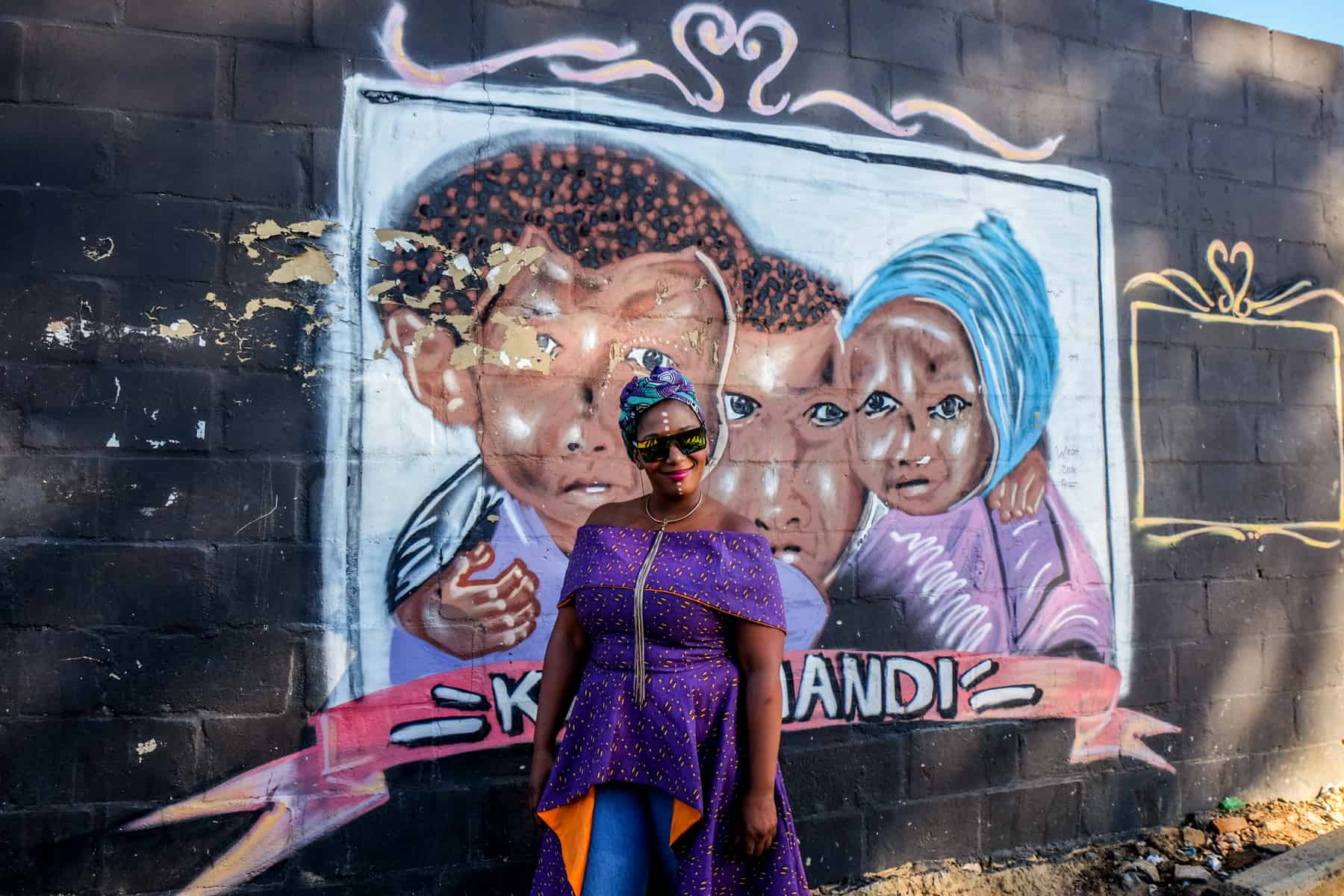 A woman in a purple dress stands in front of an art mural depicting children and with the words 'Kayamandi' - the same of the township in South Africa where she lives