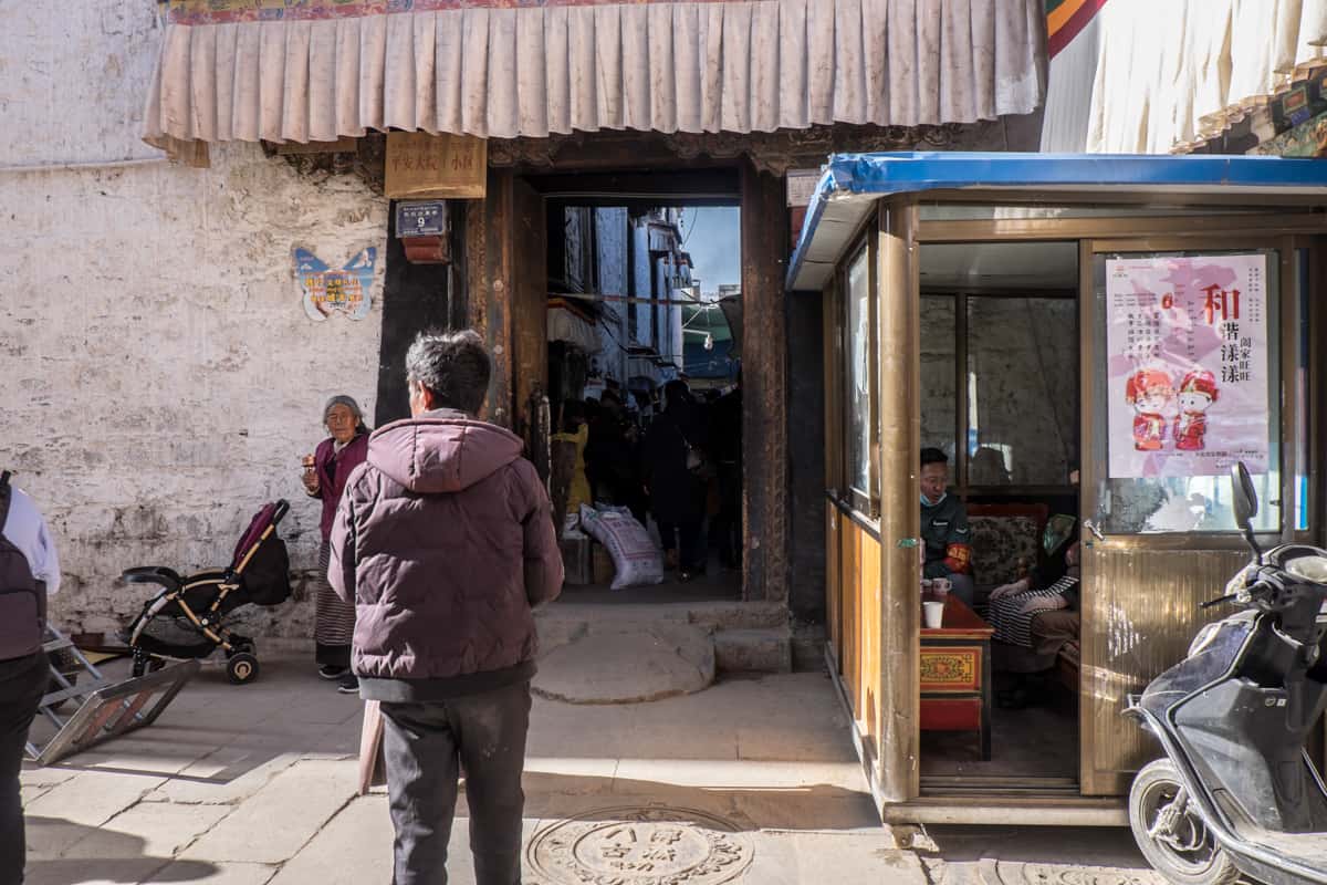 A man sits in a checkpoint box in Lhasa, Tibet while a local Tibetan man in a dark purple jacket walks by to enter an alleyway