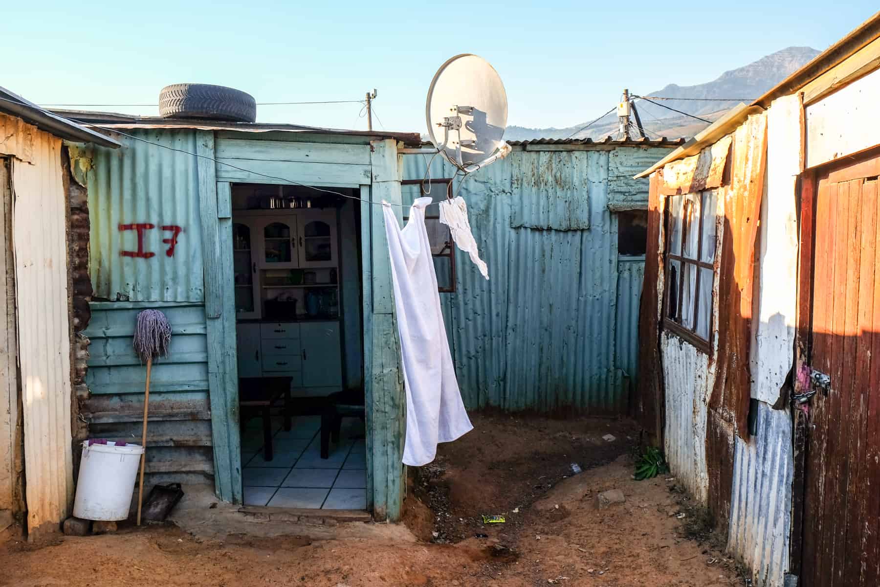 A Corrugated galvanised iron, flat-roofed house in a Township in South Africa. It is painted in a minty blue, the door is open and a white satellite dish is attached to the right corner. 