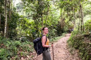 A woman with a black backpack and stick on a jungle path on the Lost City Trek.