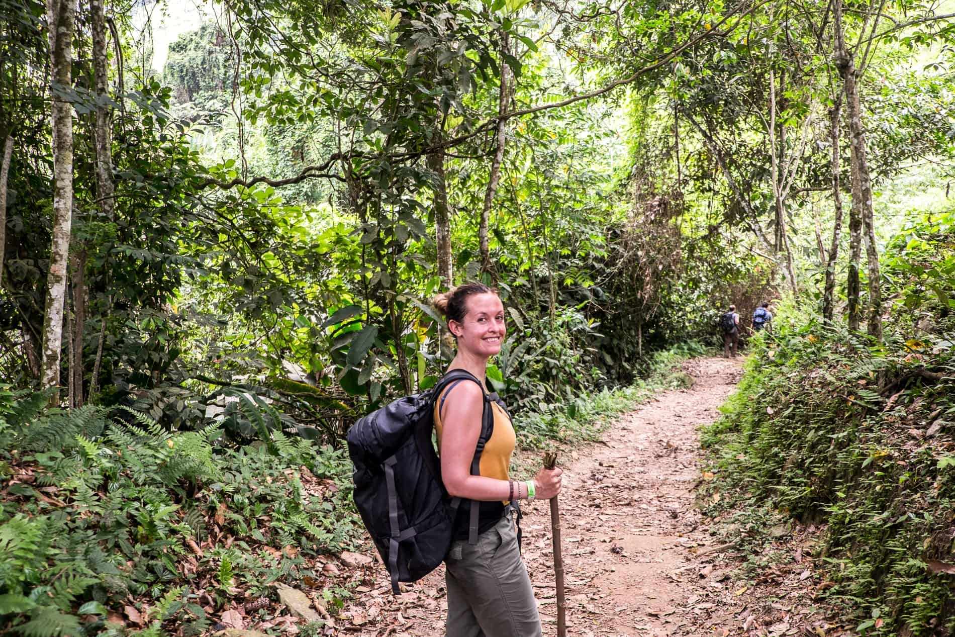 A woman with a black backpack and stick on a jungle path on the Lost City Trek. 
