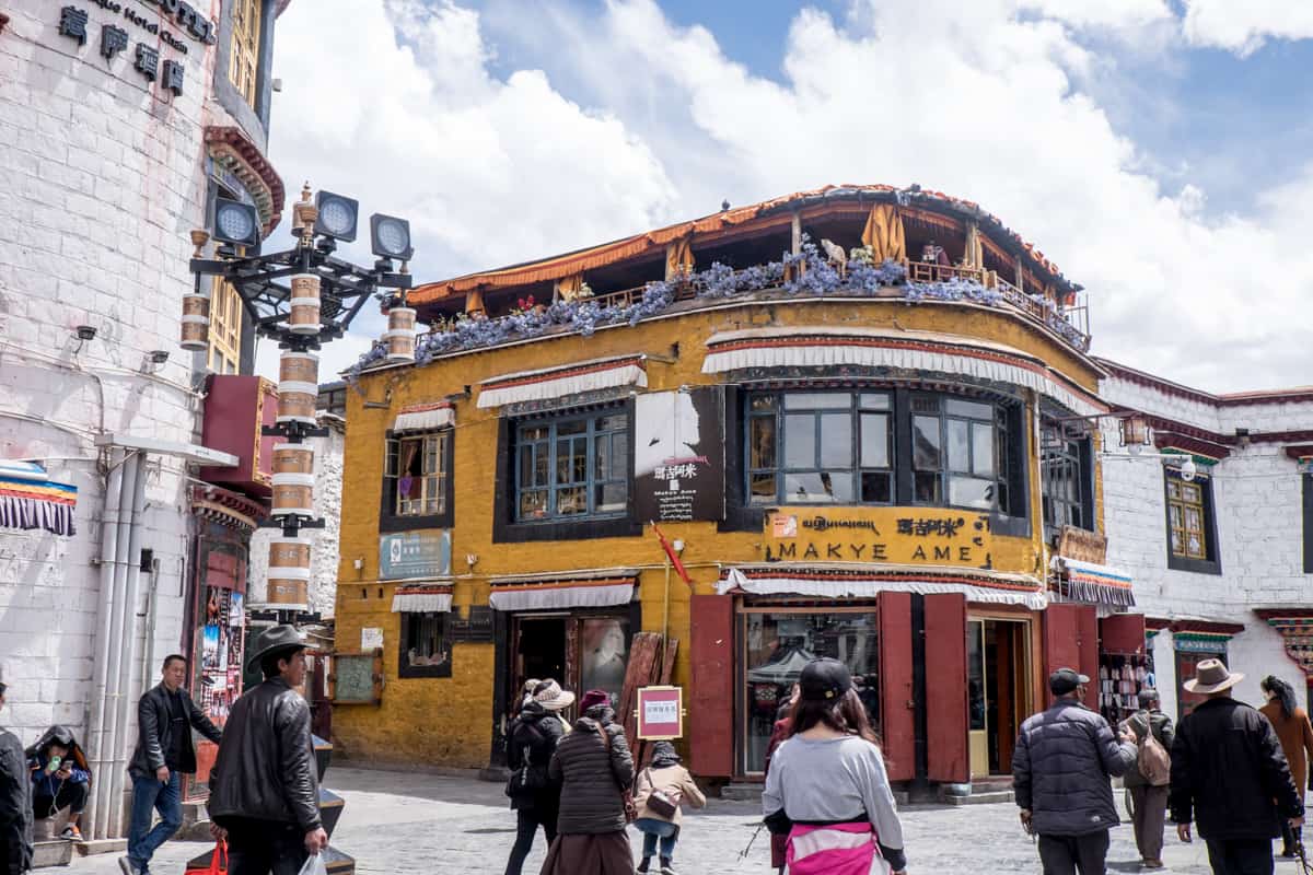 Street scene from the circular Barkhor Path in Lhasa, always filled with people walking clockwise, past the yellow Makye Ame building. A street lamp, with fitted cameras can be seen on the left.