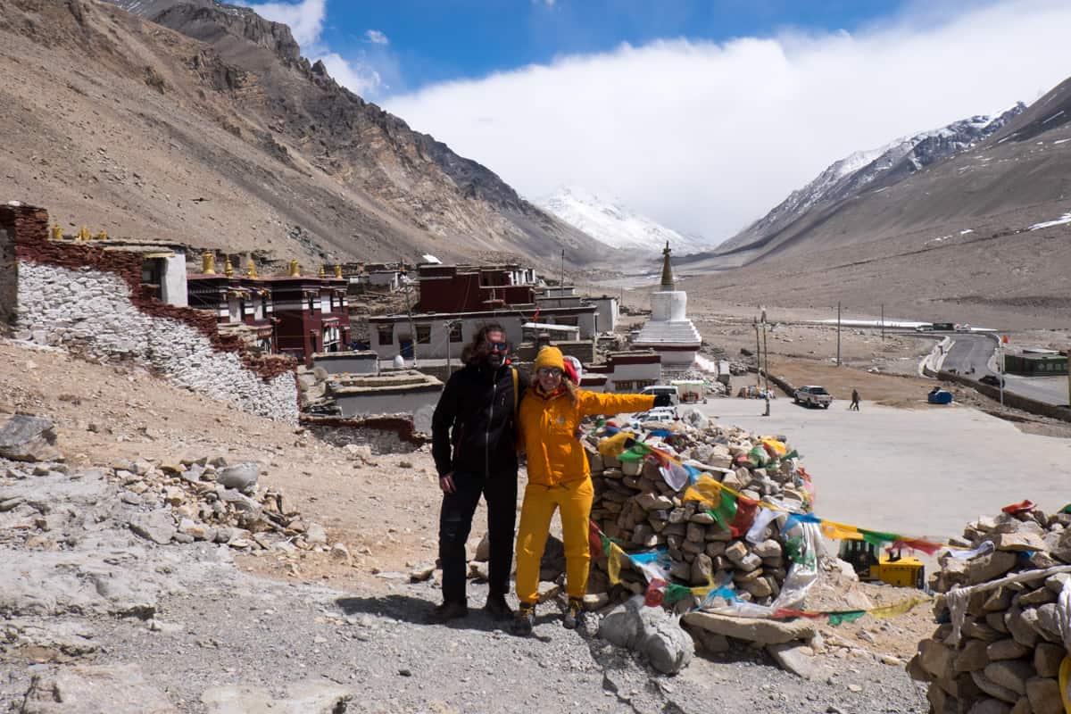 A man wearing black and a woman wearing yellow on a rocky hill with a view of Everest, beside the Rombuk Monastary at Tibet Everest Base Camp