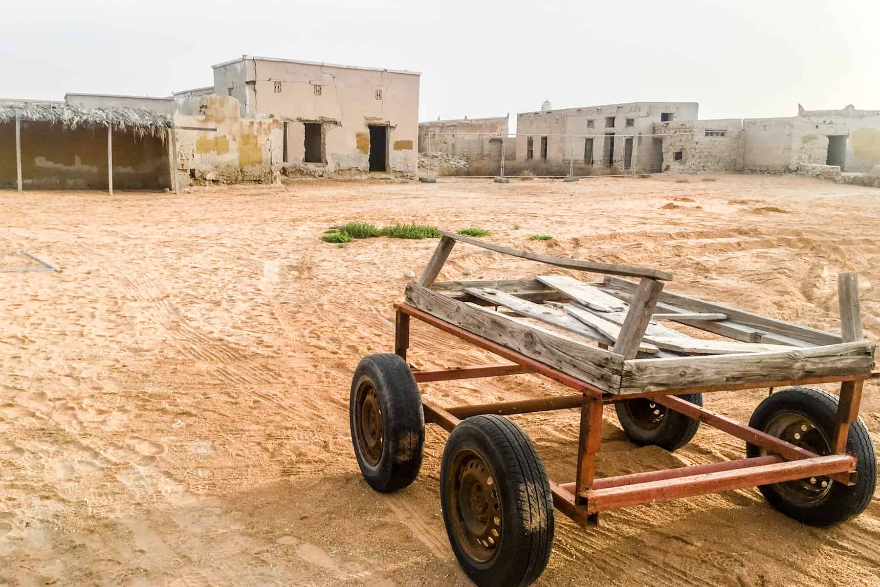A broken wooden four-wheeled cart stands abandoned on orange dust, looking towards empty shells of beige buildings in a ghost village in Ras Al Khaimah
