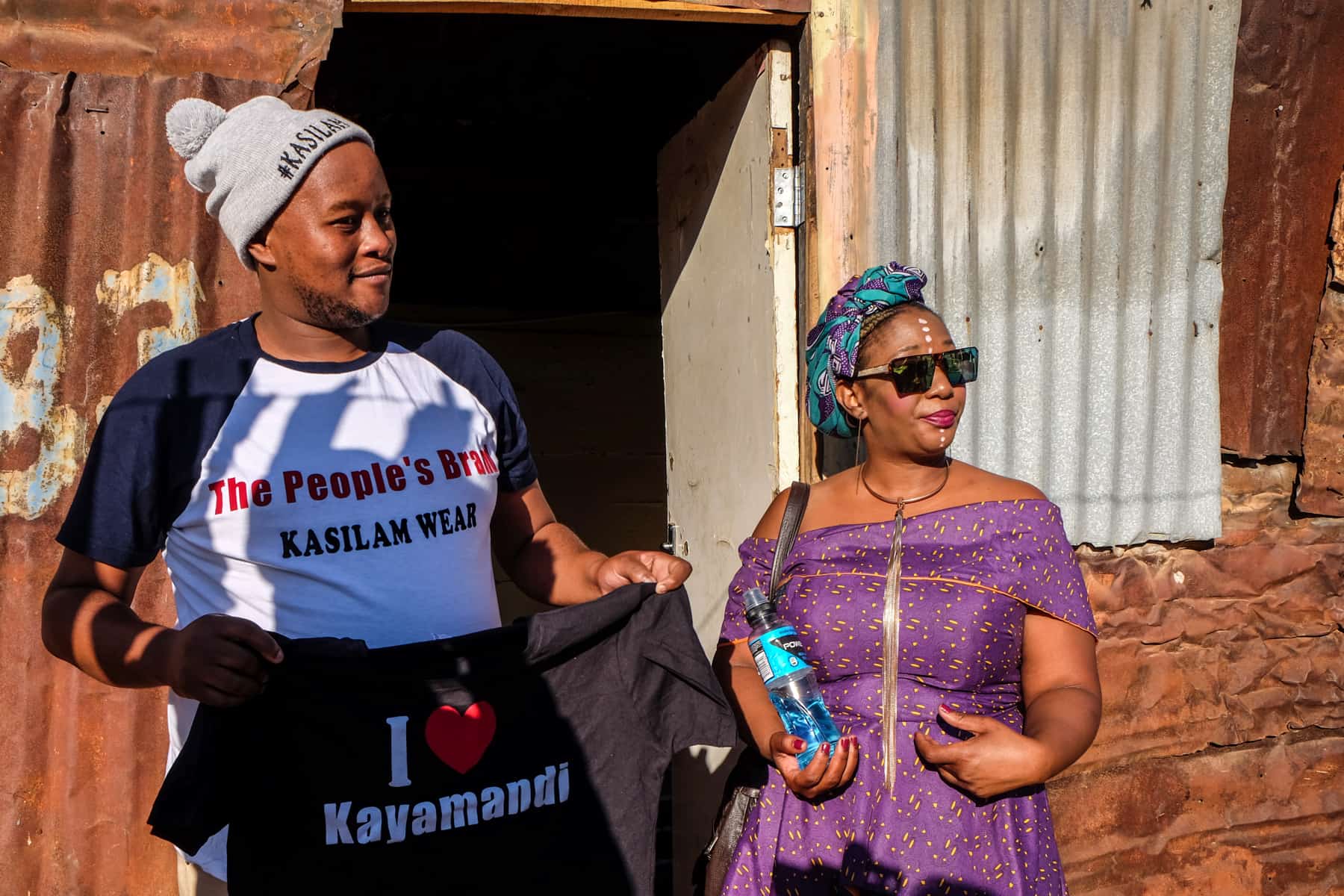 A mean wearing a grey hat holds a black t-shirt that reads 'I (heart) Kayamandi' as he stands next to a woman in a purple dress outside of a clay mud and tin shack house