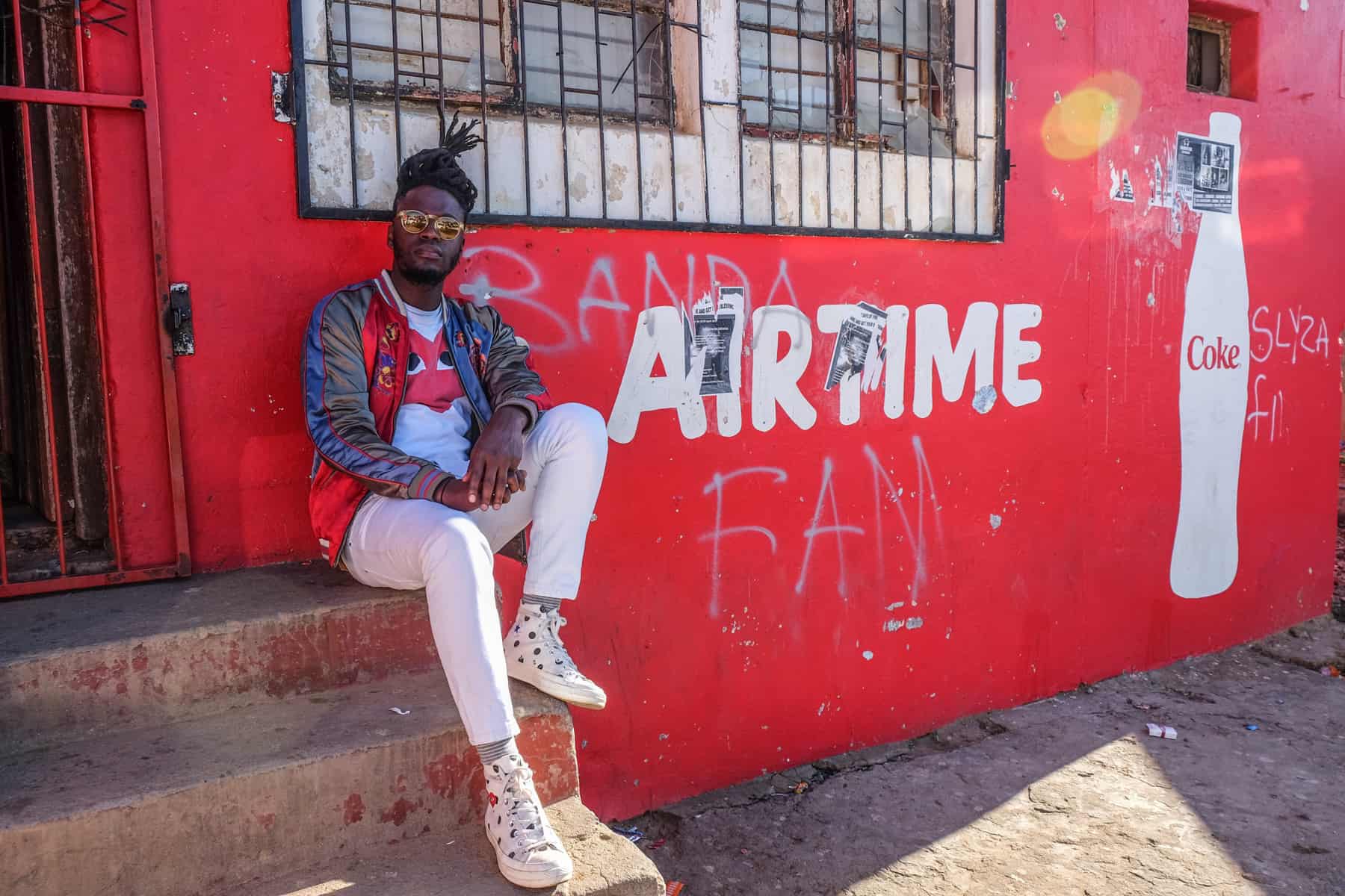 A man dressed in white sits on the steps of a red painted convenience store in a Township in South Africa