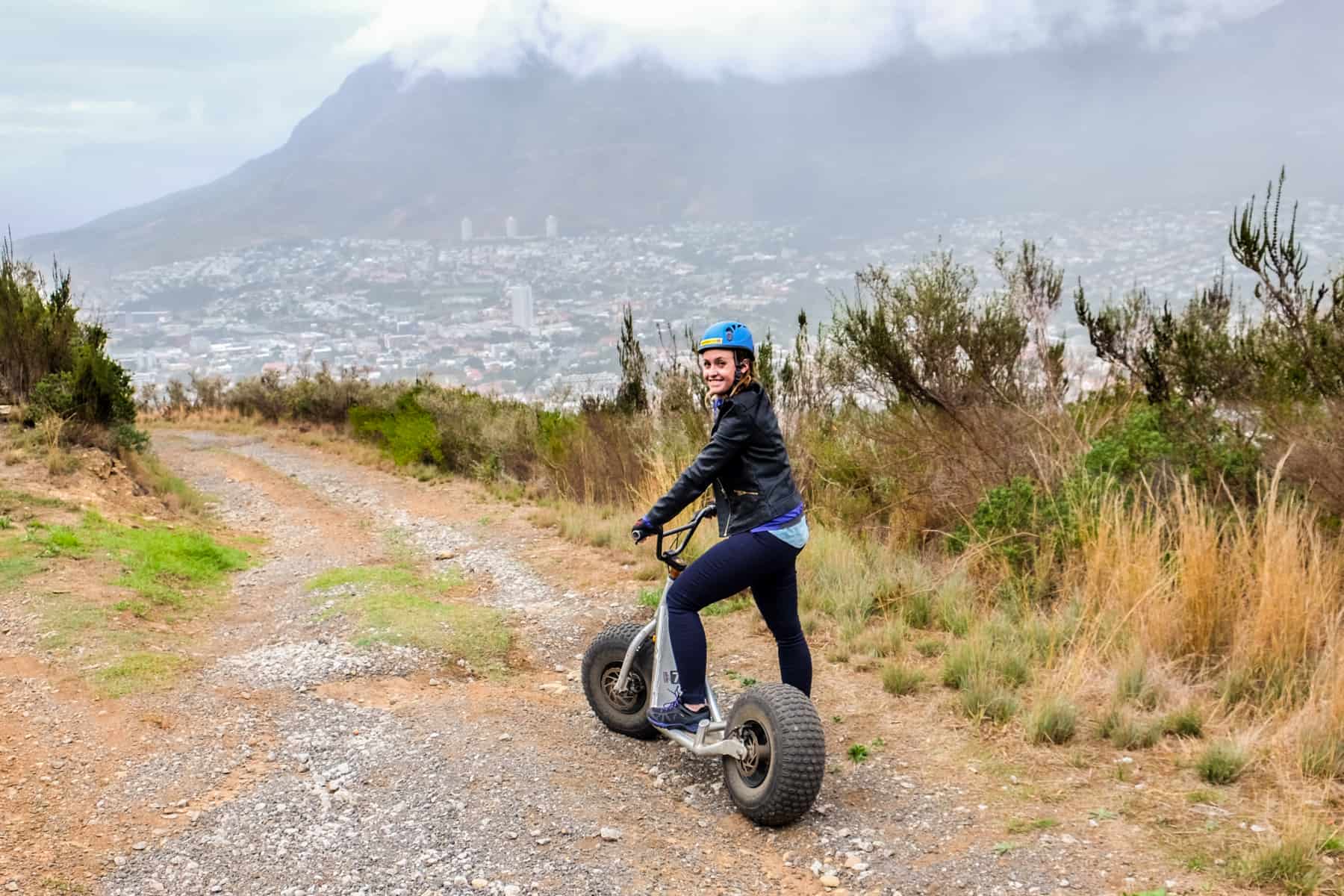 A woman wearing black rides a scooter down the yellow, gravel slopes of Table Mountain in Cape Town. On the right you can see the far end of the mountain, covered by clouds.