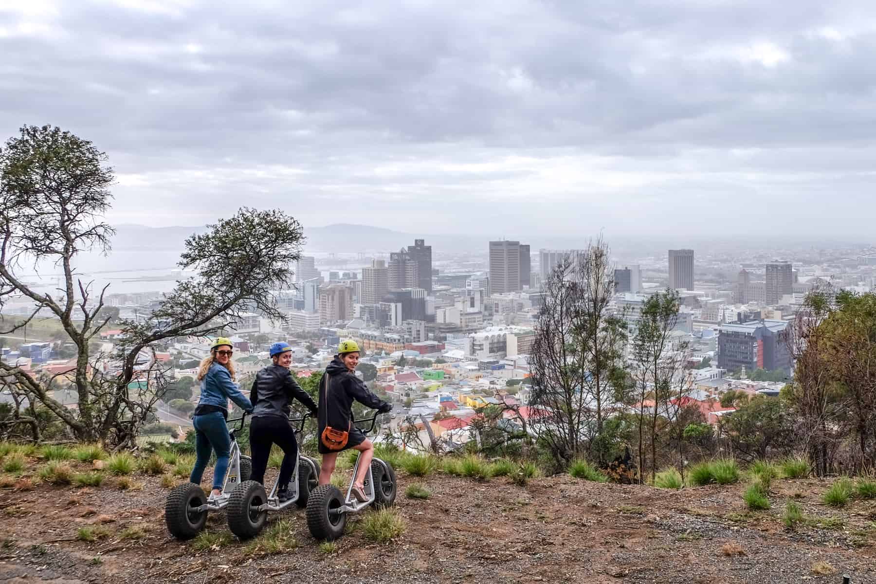 Three women on scooters look towards the camera. In front of them is the high rises and city spread of Cape Town, South Africa