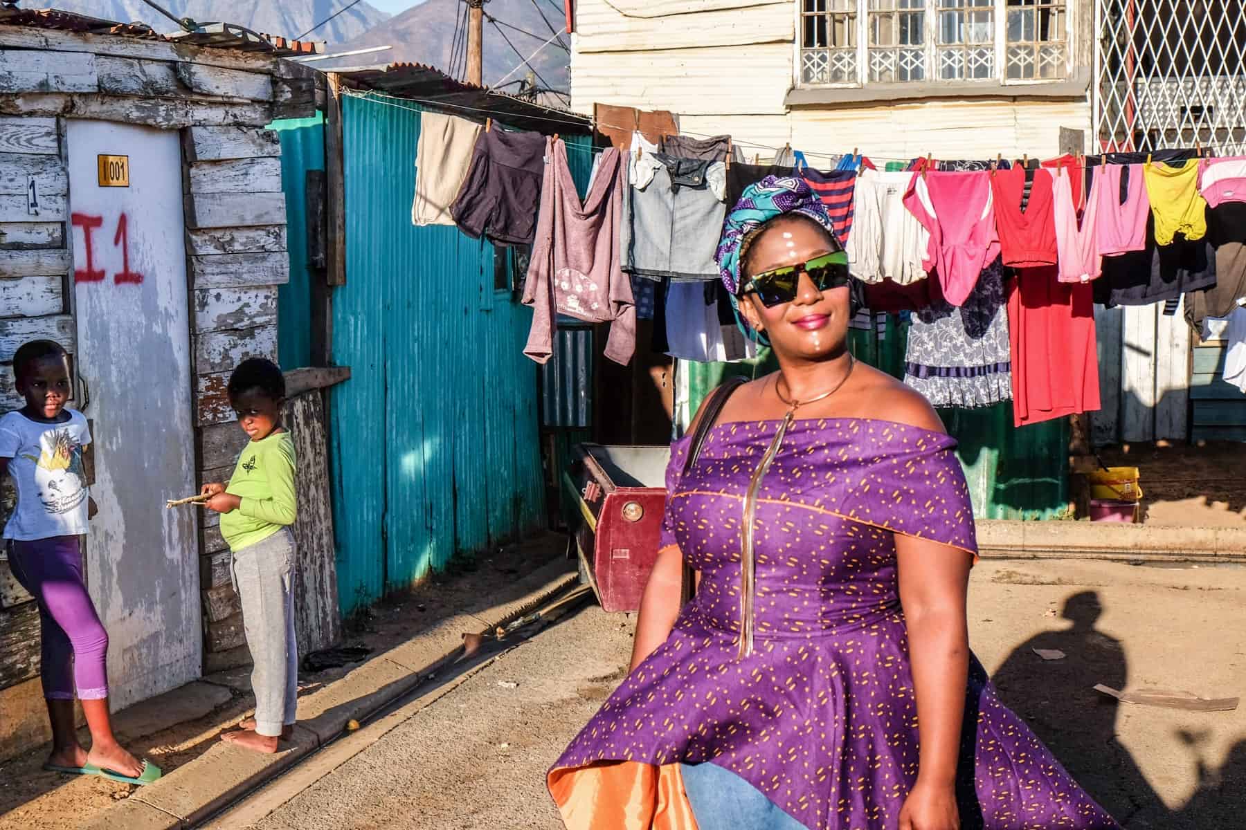 A local woman, Thembi, dressed in purple, takes visitors on a tour of her home township Kayamandi. Behind her is a row of washing, two children and some iron huts.
