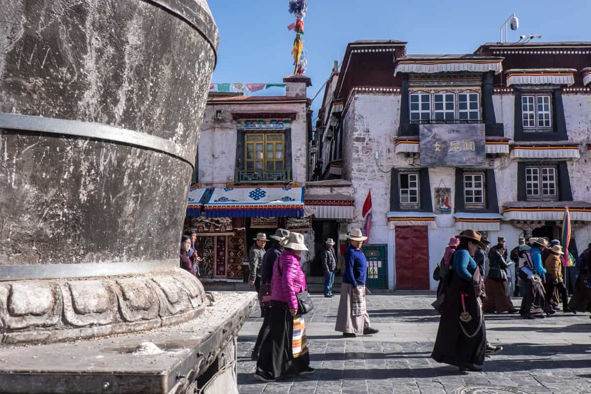 Tibetans in Lhasa walking in a clockwise direction around the sacred Kora path called The Barkhor in Lhasa, Tibet. They pass large stone structures and white buildings with red window frames.