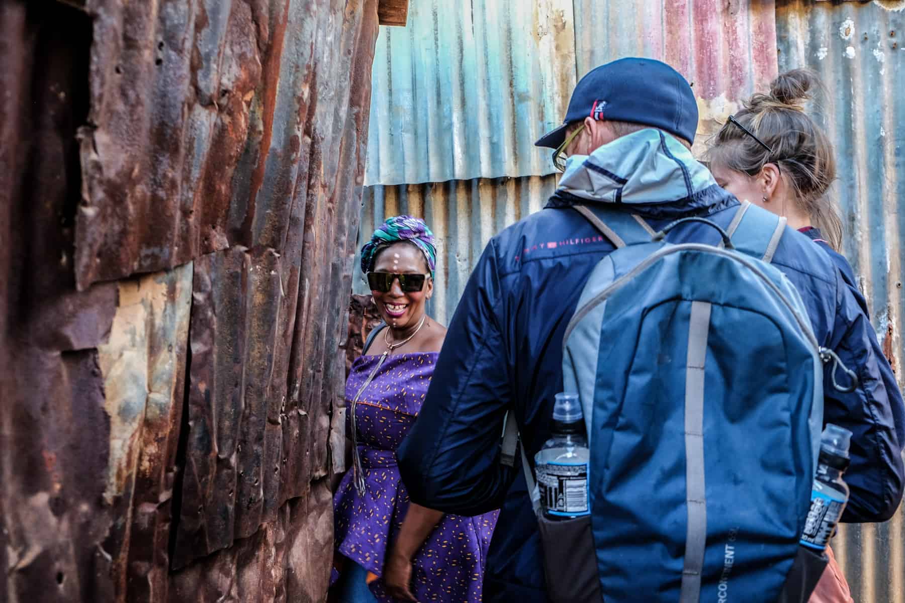 A woman in a purple dress guides a woman in blue through a narrow corridor between houses made of corrugated iron, in a township in South Africa.