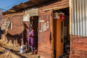 A local woman in a purple dress stands in the doorway of a clay coloured one-floor housing unit in a township. She is guiding a tour group through her home in Kayamandi