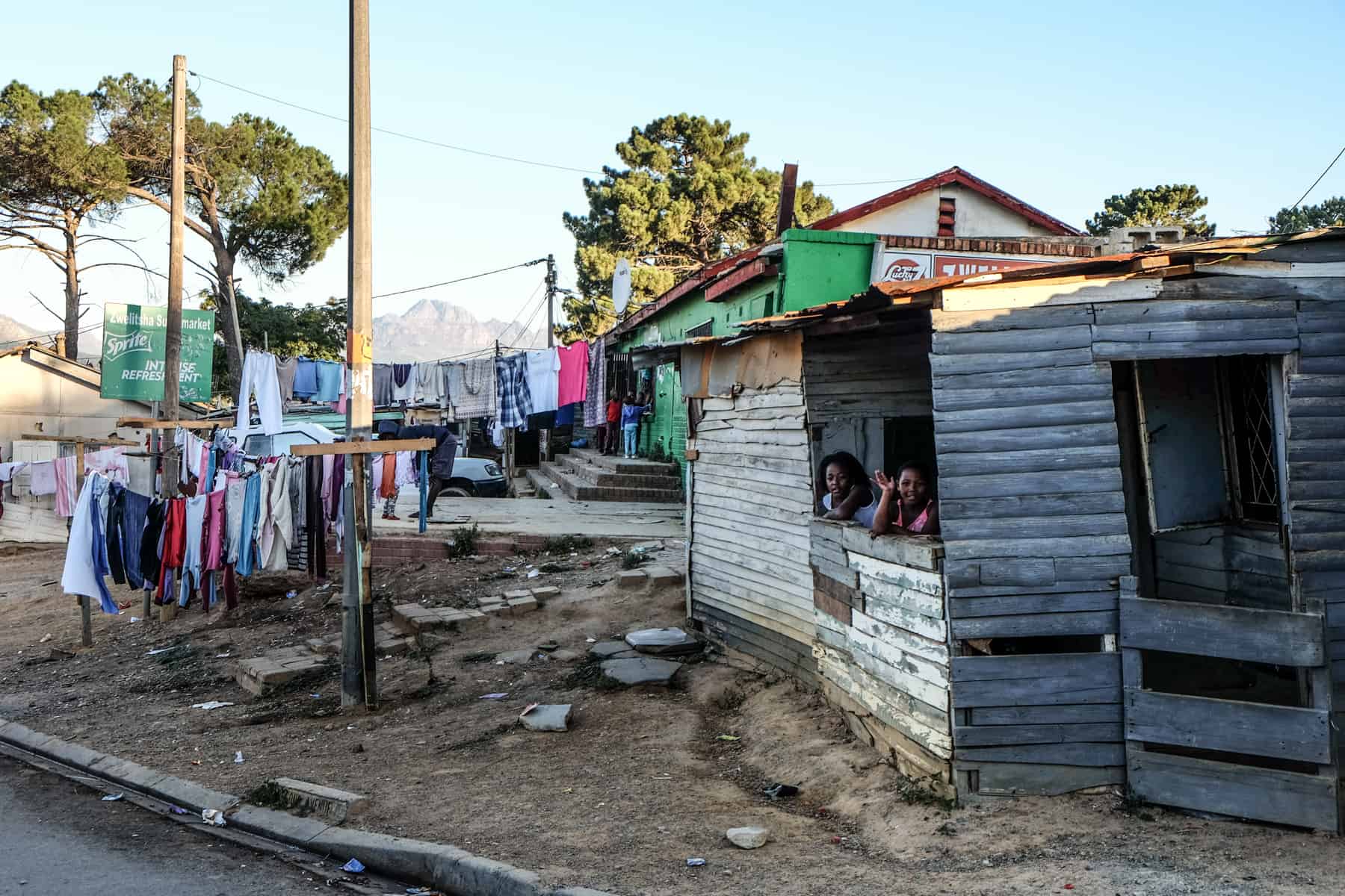 Two girls waves from their slanted wooden house in Kayamandi township in South Africa next to a dirt path with a washing line