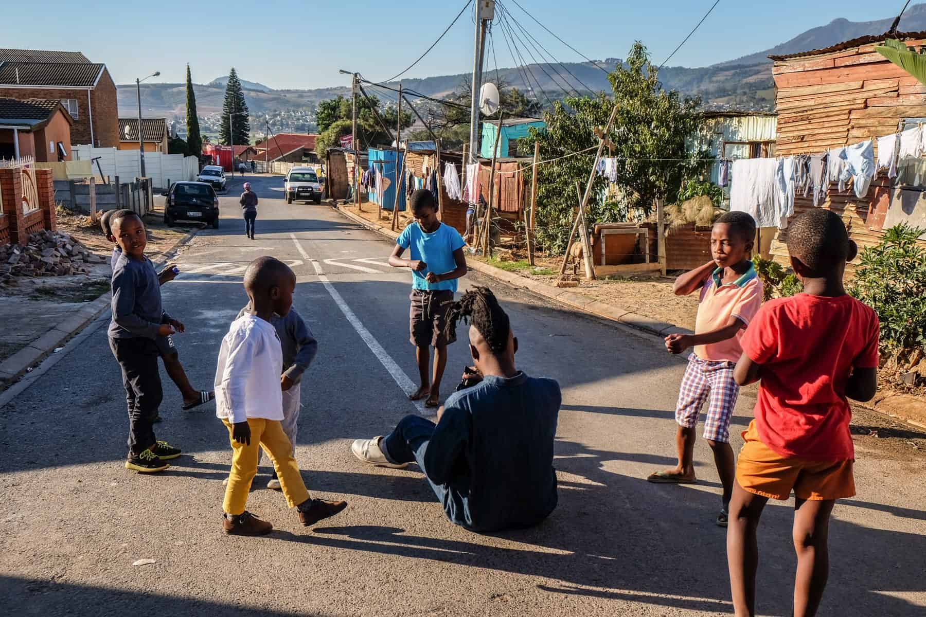 A man in a blue jean jumpsuit sits on the paved road in a South African township surrounded by five children. In the background are some houses, and hills in the far distance
